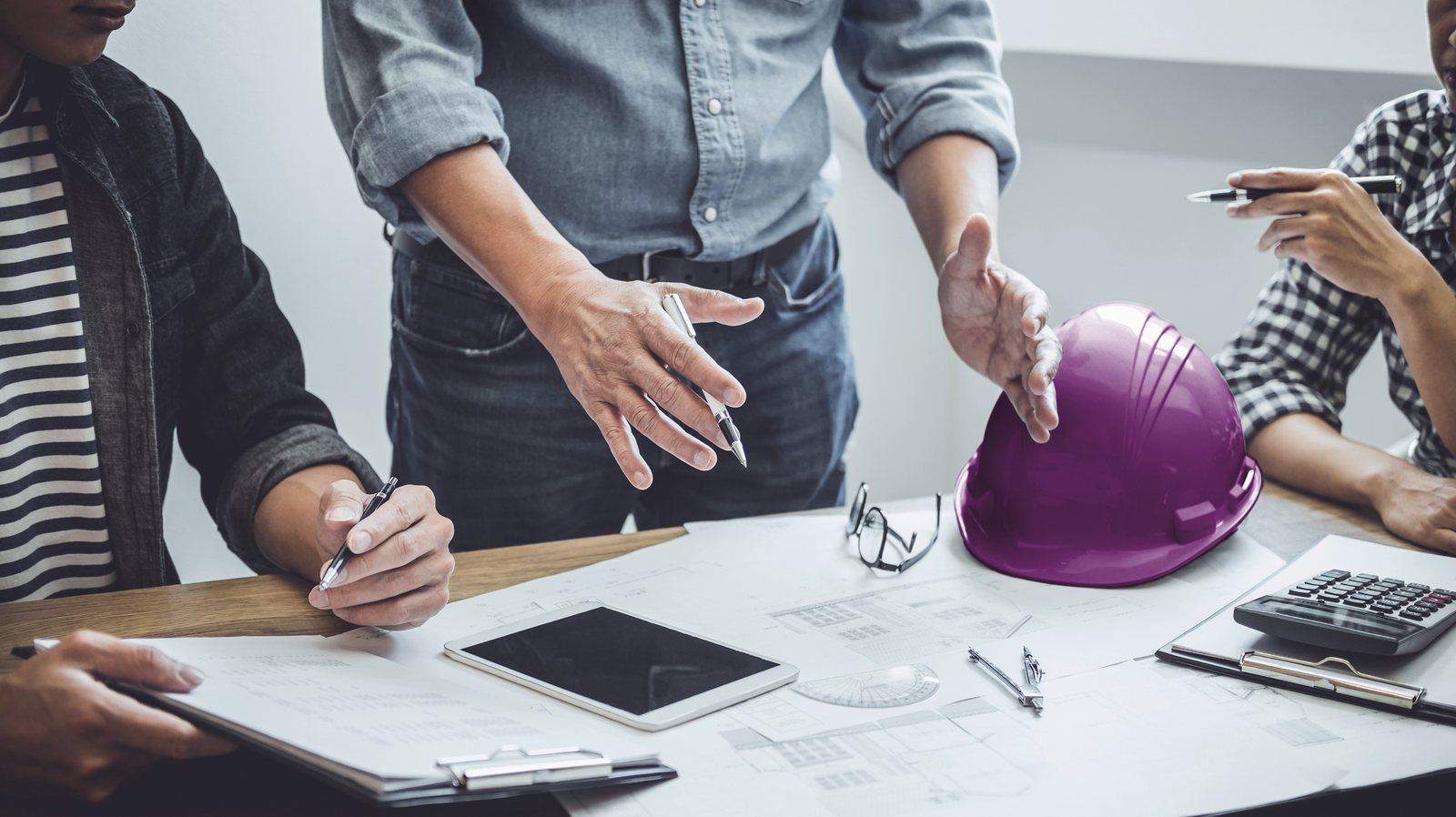 Table de r&eacute;union vue de haut&nbsp;: plans, tablette, calculatrice et compas, casque de chantier magenta au centre.