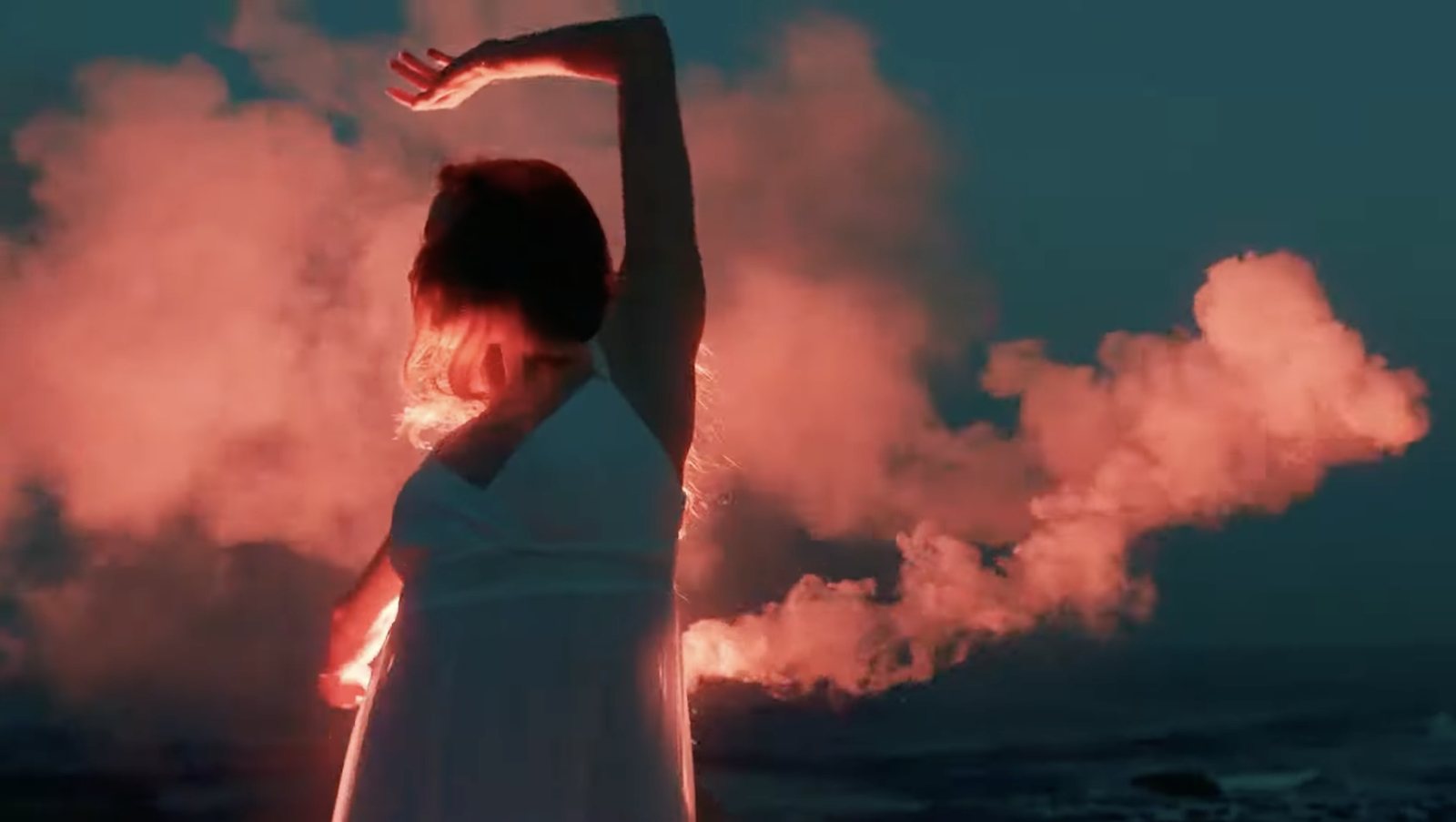 Plan du film Stelliant Expertise&nbsp;: danseuse en robe blanche sur un rivage, travers&eacute;e de fum&eacute;e orang&eacute;e.